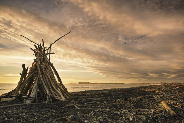 Tell The Sun Not To Shine - A driftwood teepee on the beach with a sunrise view towards Napier, New Zealand.